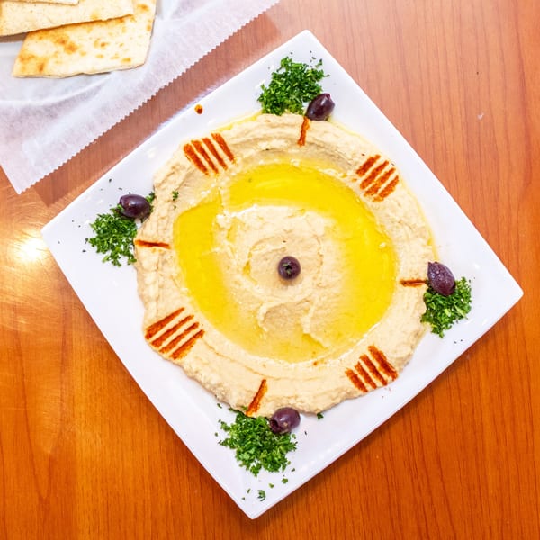 Hummus with olive oil, paprika, parsley, and olives, served with pita bread on a wooden table.