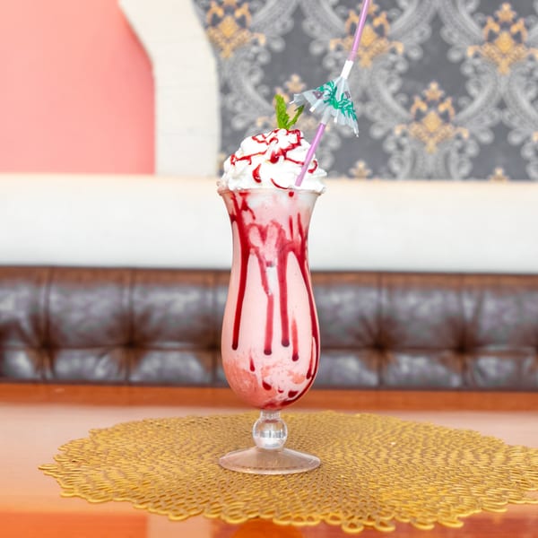 A tall glass of strawberry milkshake with whipped cream, red syrup, mint, and a paper umbrella.